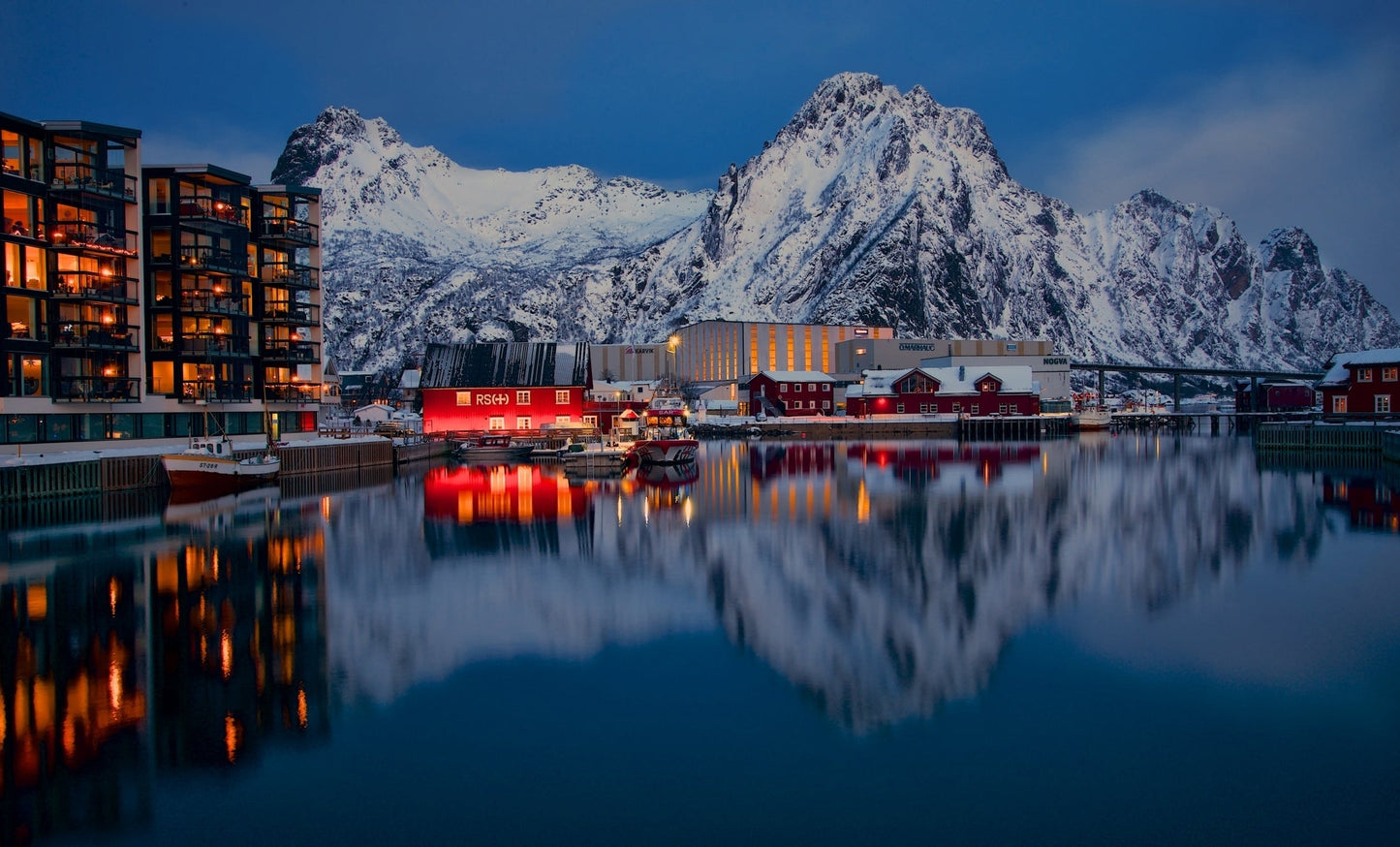 Lake in Lofoten Norway with buildings and snow covered mountains in the background.