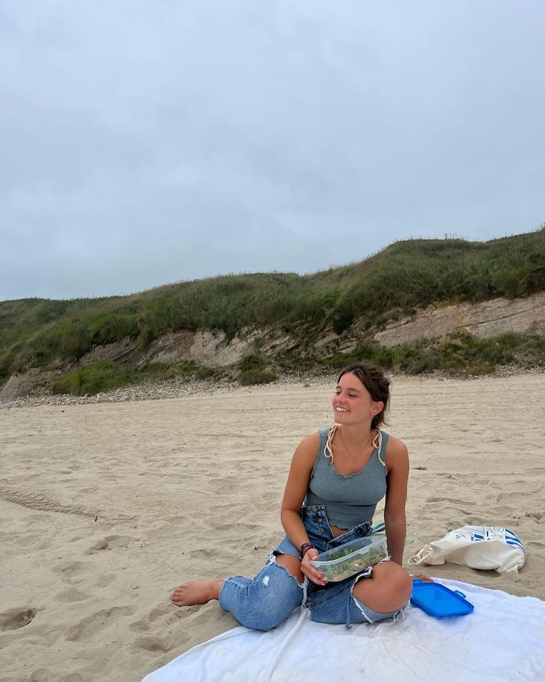 Trip leader Miriam sitting on a blanket in the sand with a Tupperware in hand.