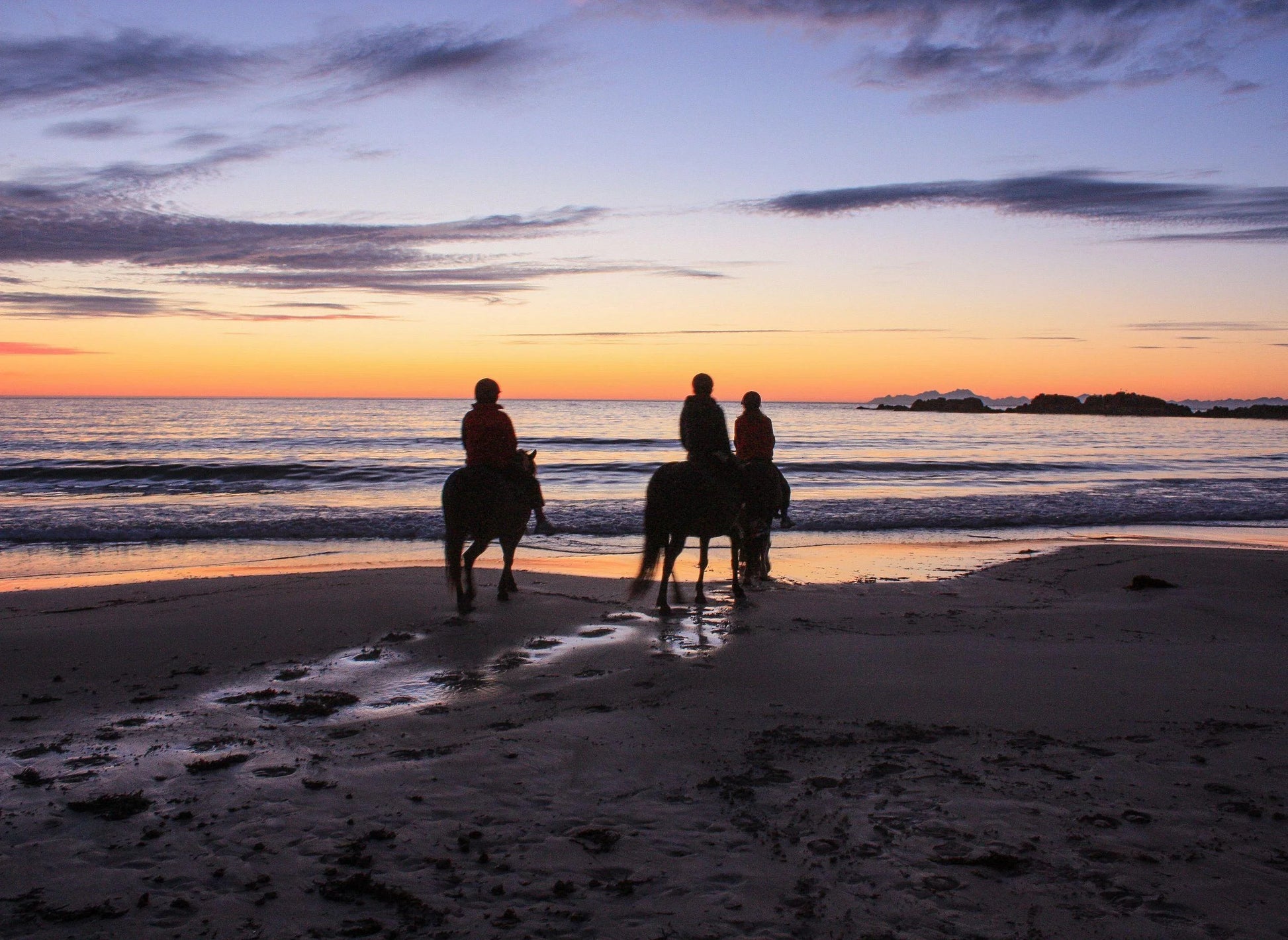 Three small group trip participants riding horses along the beach at sunset in Lofoten Norway.
