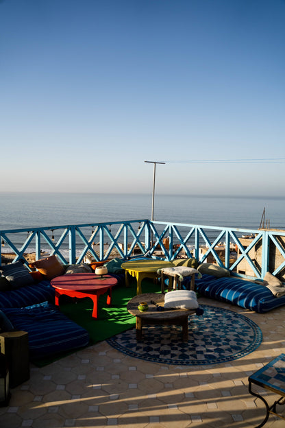 Oceanview Taghazout hostel outdoor seating area next to the beach with tables and cushions.
